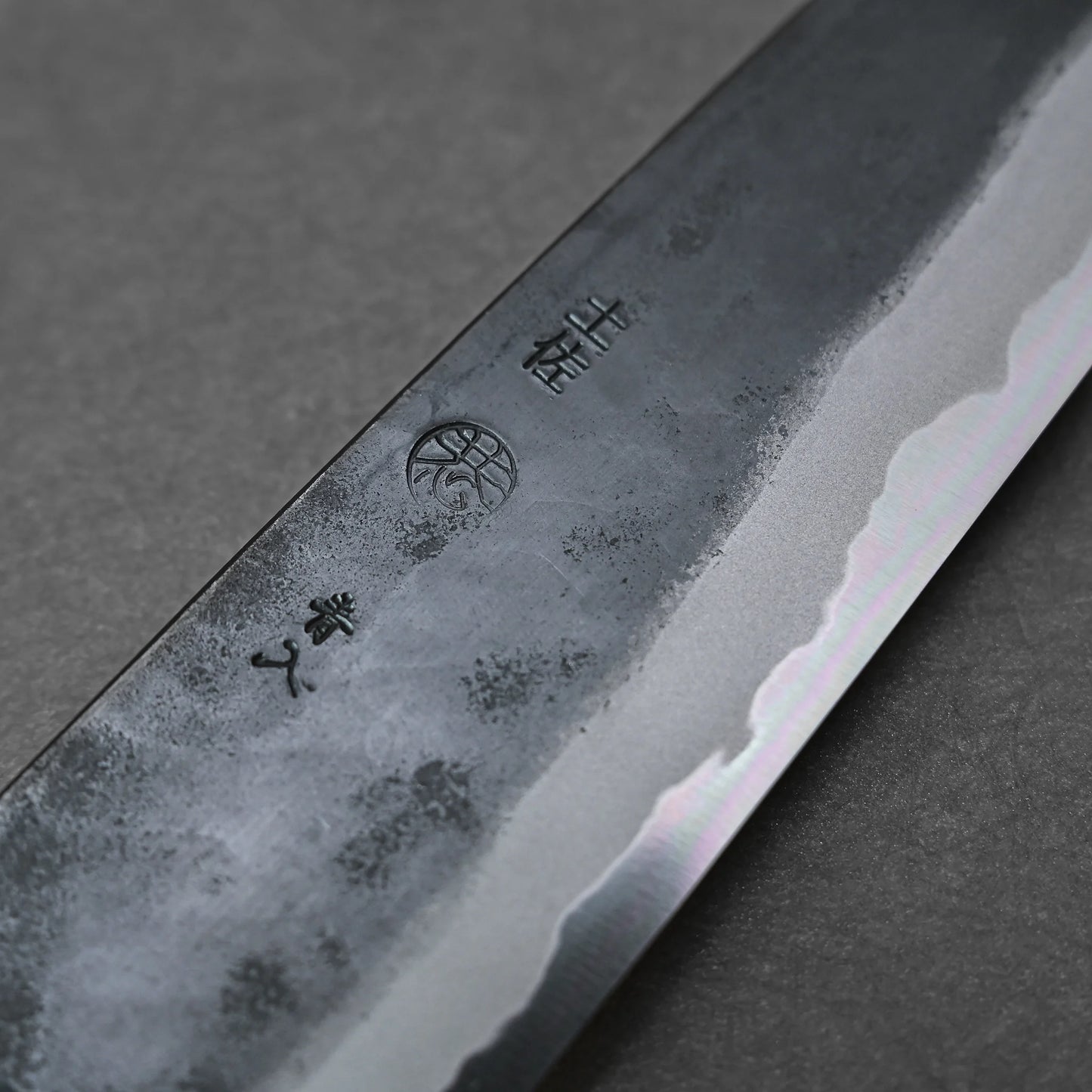 Close-up of the Kyohei Shindo aogami#2 kurouchi gyuto 240mm blade, showing hamon pattern, blacksmith marks, engraved kanji, and a circular emblem on Aogami#2 carbon steel with a kurouchi finish against a dark background.