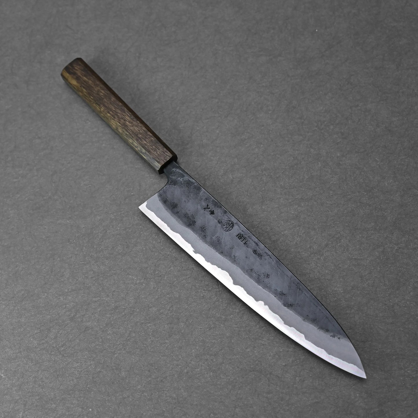 The Kyohei Shindo aogami#2 kurouchi gyuto 240mm features a dark wooden handle and a two-tone Aogami#2 carbon steel blade with a hamon line, shown on a gray textured surface.