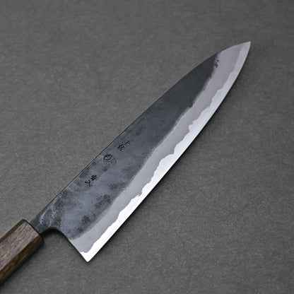The Kyohei Shindo aogami#2 kurouchi gyuto 240mm features a wooden handle and sharp, polished carbon steel blade with Japanese characters near the handle, showcasing its expert craftsmanship on a dark gray surface.