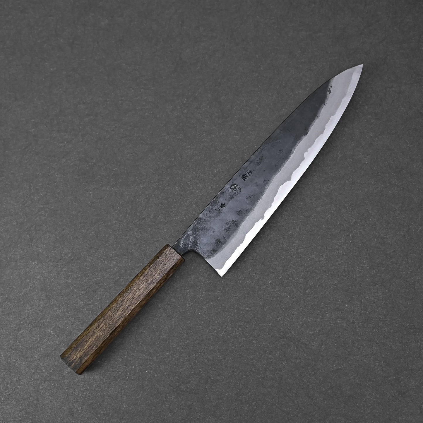 The Kyohei Shindo aogami#2 kurouchi gyuto 240mm features a dark wooden handle and an Aogami#2 carbon steel blade with a kurouchi finish, displayed on a gray textured surface.