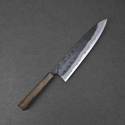 The Kyohei Shindo aogami#2 kurouchi gyuto 240mm features a dark wooden handle and an Aogami#2 carbon steel blade with a kurouchi finish, displayed on a gray textured surface.