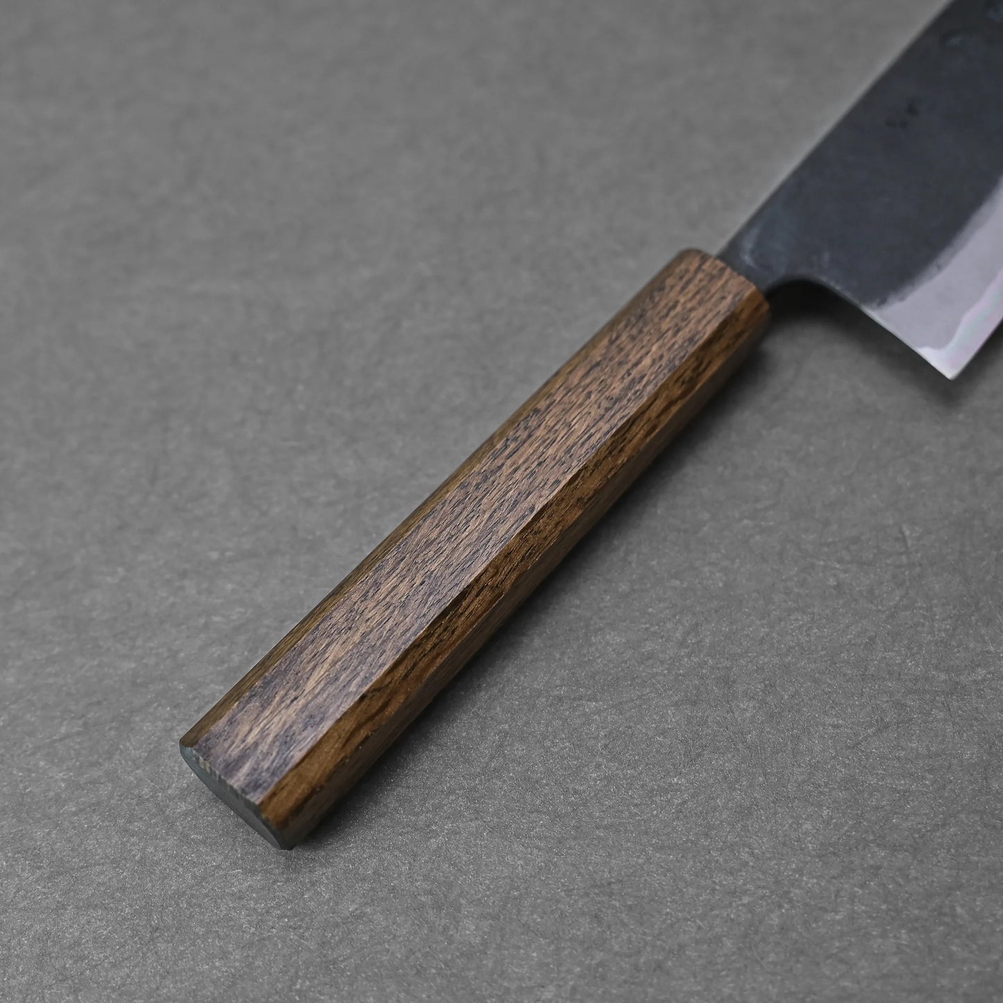 A close-up of the Kyohei Shindo aogami#2 kurouchi gyuto 240mm, featuring a straight, dark wooden handle and partially visible carbon steel blade, resting on a gray textured surface.