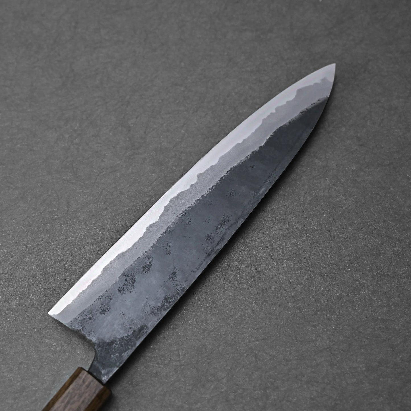 A close-up of the Kyohei Shindo aogami#2 kurouchi gyuto 240mm, showing its wavy hamon on the Aogami#2 carbon steel blade, laying on a dark gray textured surface.