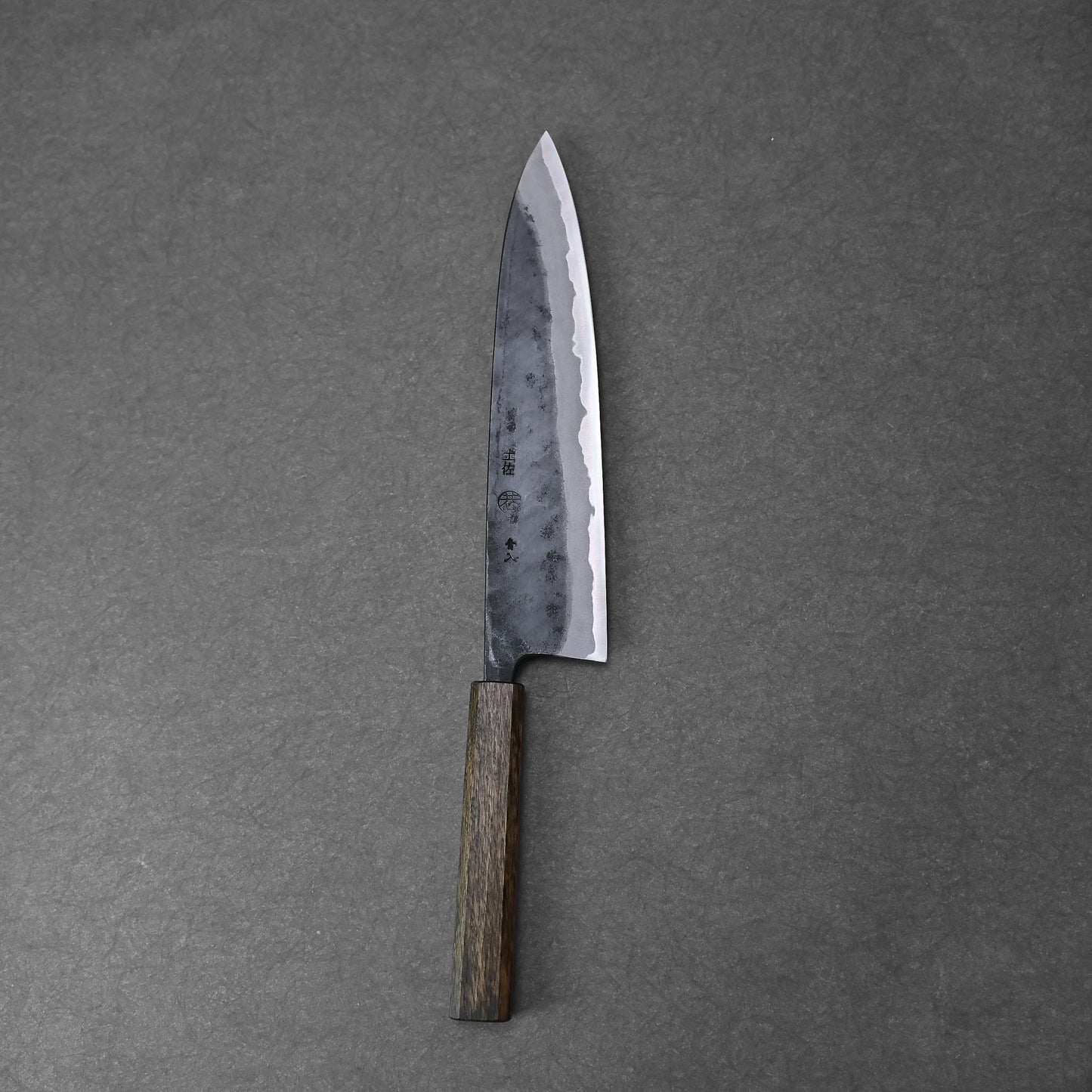 The Kyohei Shindo aogami#2 kurouchi gyuto 240mm, featuring a dark wooden handle and a sharp, textured Aogami#2 carbon steel blade, rests on a dark gray surface.