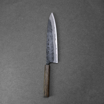 The Kyohei Shindo aogami#2 kurouchi gyuto 240mm, featuring a dark wooden handle and a sharp, textured Aogami#2 carbon steel blade, rests on a dark gray surface.