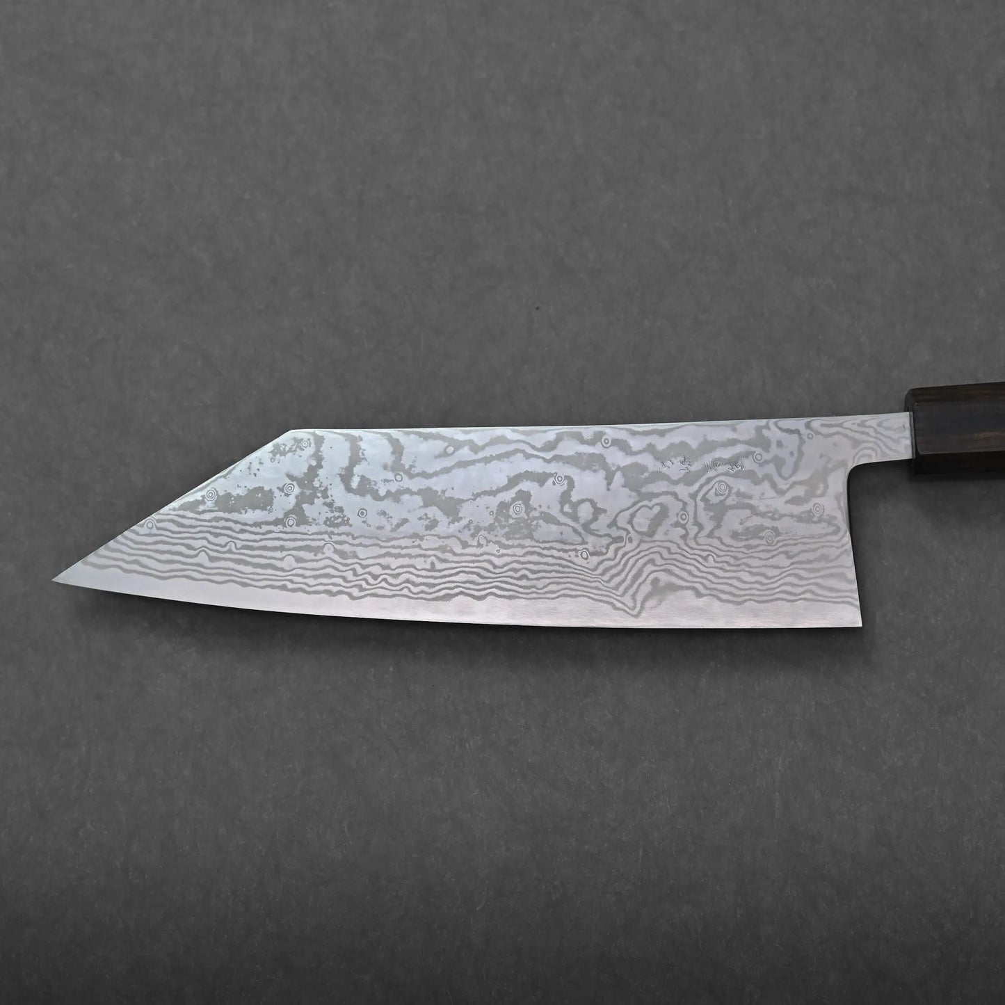 A close-up of a chefs knife with a distinctive, wavy Damascus steel pattern on the blade—possibly a Miyazaki bunka—is shown against a plain, dark gray background. The wooden handle is partially visible on the right side.