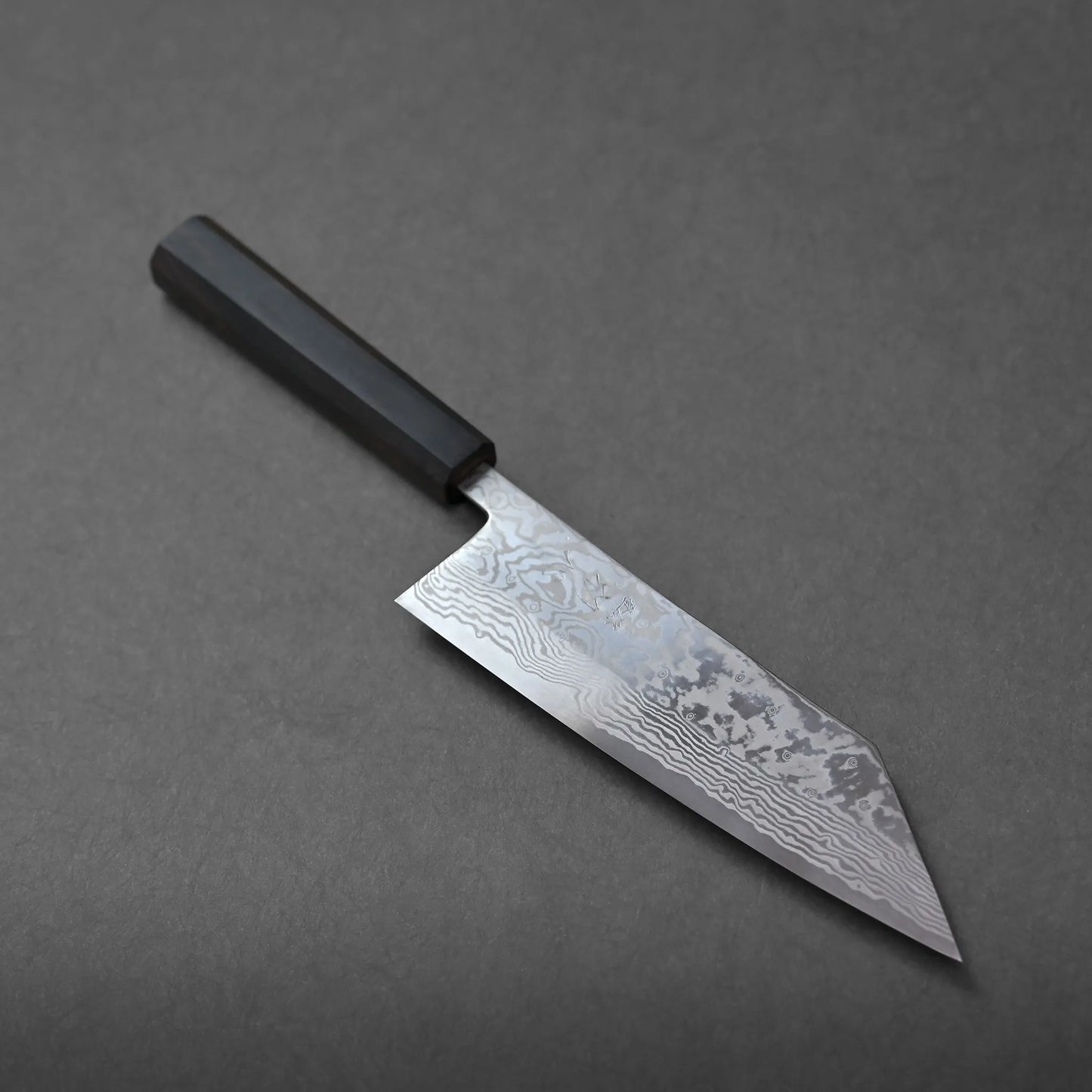 A chef’s knife with a dark handle and a patterned blade is displayed on a gray surface. The blade features visible wavy lines, characteristic of the Miyazaki ATS34 bunka style.