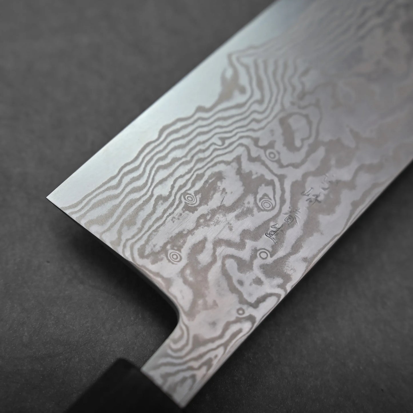 Close-up of a Miyazaki ATS34 bunka knife blade with visible Damascus steel patterning, featuring intricate, wavy lines and swirls on the metal surface against a dark background.