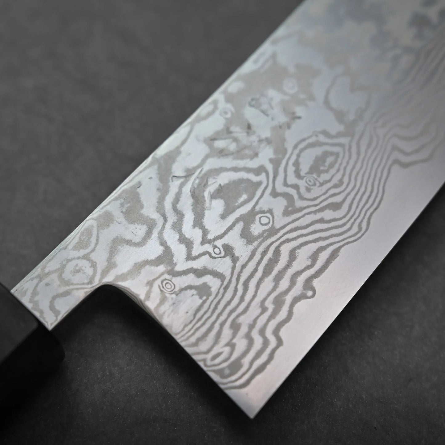 Close-up of a Miyazaki ATS34 bunka Damascus steel knife blade, displaying wavy and swirling patterns on the metal. A black handle is partially visible on the left, with the knife resting against a dark background.