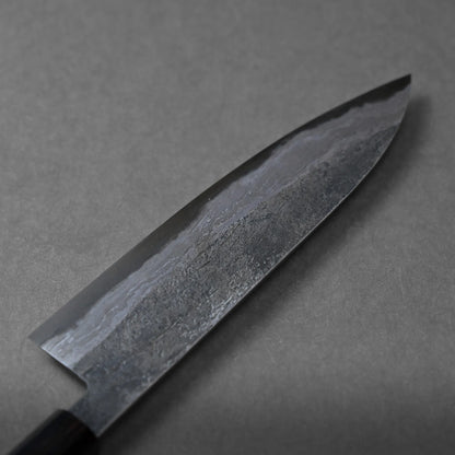 A close-up view of a dark, textured chef’s knife blade with a subtle wave pattern and hammered finish, set against a plain gray background.