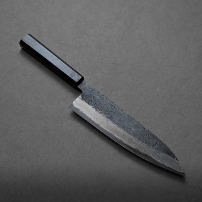 A Japanese-style chef’s knife with a dark wooden handle and a textured, partially polished blade rests on a plain grey surface, showcasing its artisanal craftsmanship.