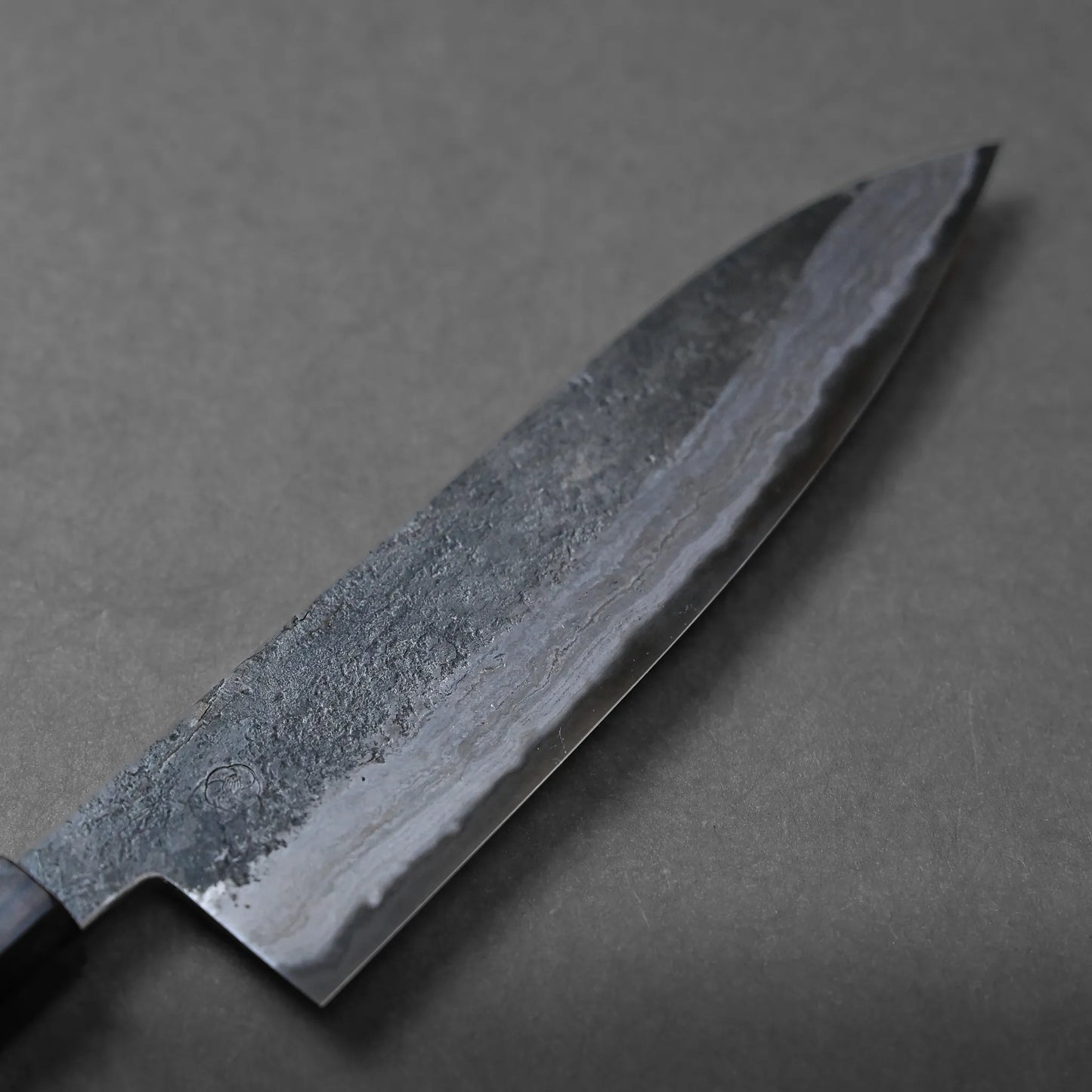 A close-up of a chef’s knife with a dark, textured blade and a smooth cutting edge, placed on a plain gray surface, highlighting its unique craftsmanship.