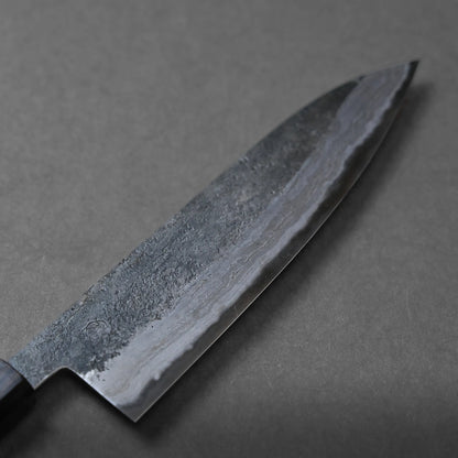 A close-up of a chef’s knife with a dark, textured blade and a smooth cutting edge, placed on a plain gray surface, highlighting its unique craftsmanship.