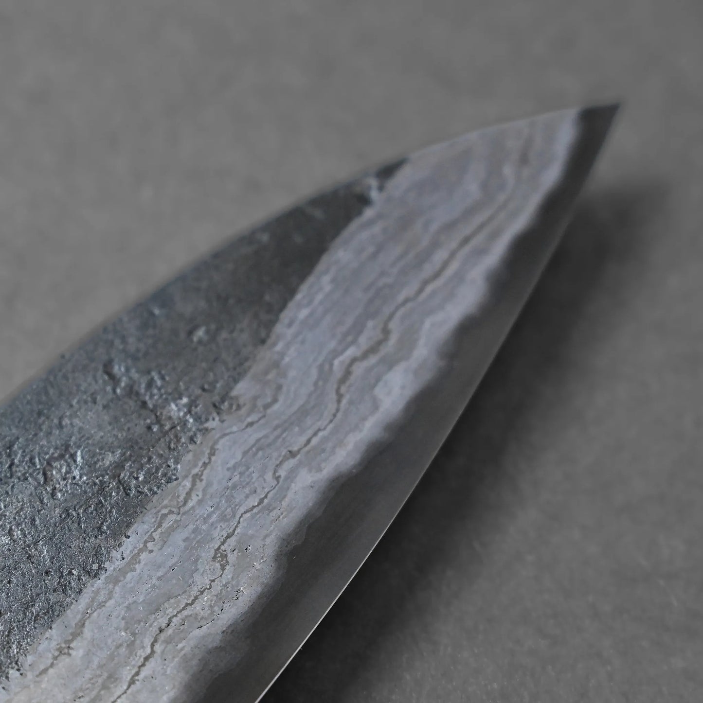 Close-up of a knife blade revealing a wavy, patterned texture along the metal edge, highlighting a classic Damascus steel design against a gray background.