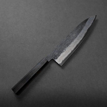A Miyazaki kurouchi rentetsu aogami#1 gyuto 220mm with a dark wooden handle and a broad, slightly curved blade rests elegantly on a plain, dark surface.