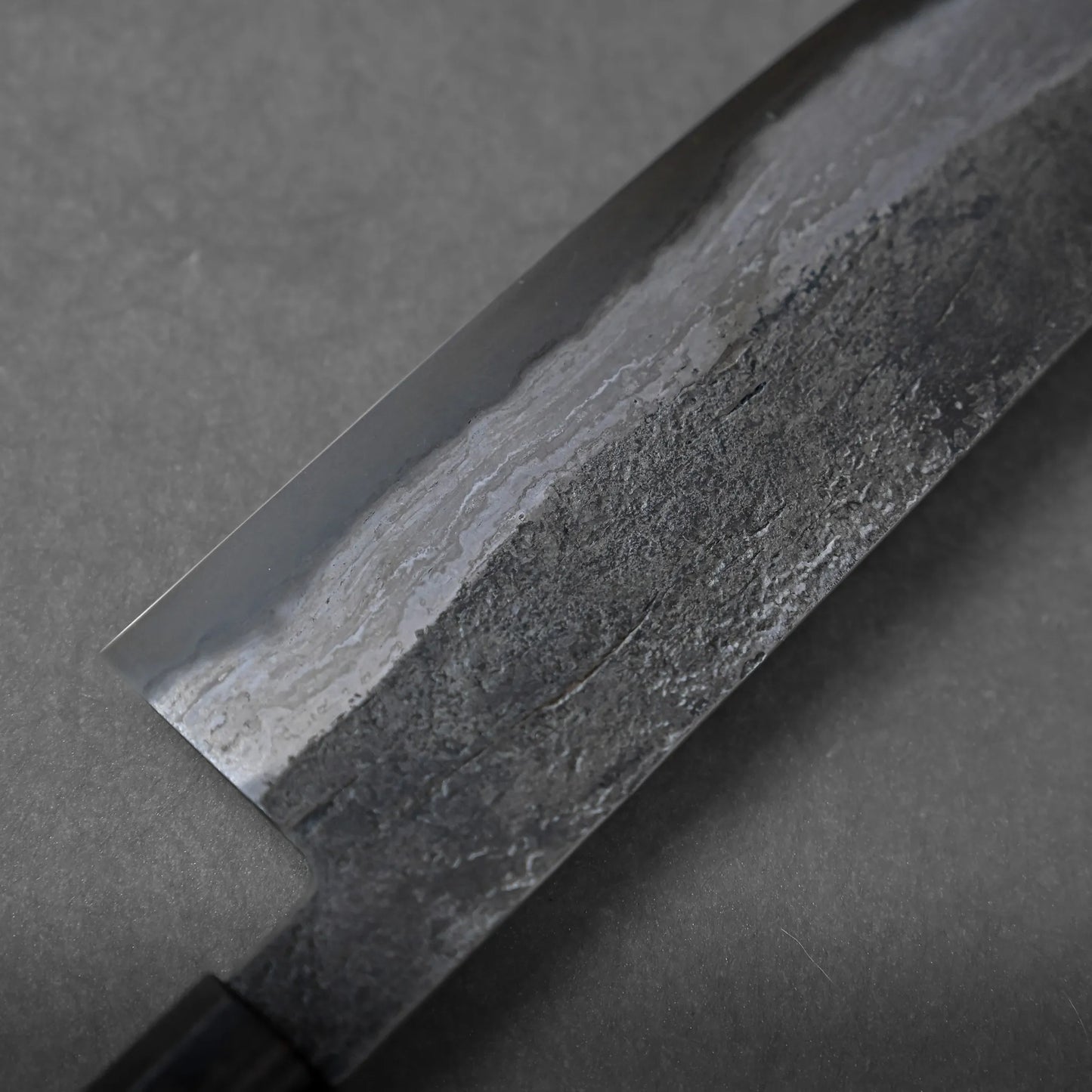 Close-up of a Japanese-style kitchen knife with a textured, partially polished blade and a dark handle, resting on a gray surface. The blade displays traditional craftsmanship with contrasting smooth and rough finishes.