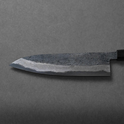 A close-up of a Japanese-style kitchen knife with a textured, dark blade and wavy Damascus pattern near the edge, lying on a gray surface.