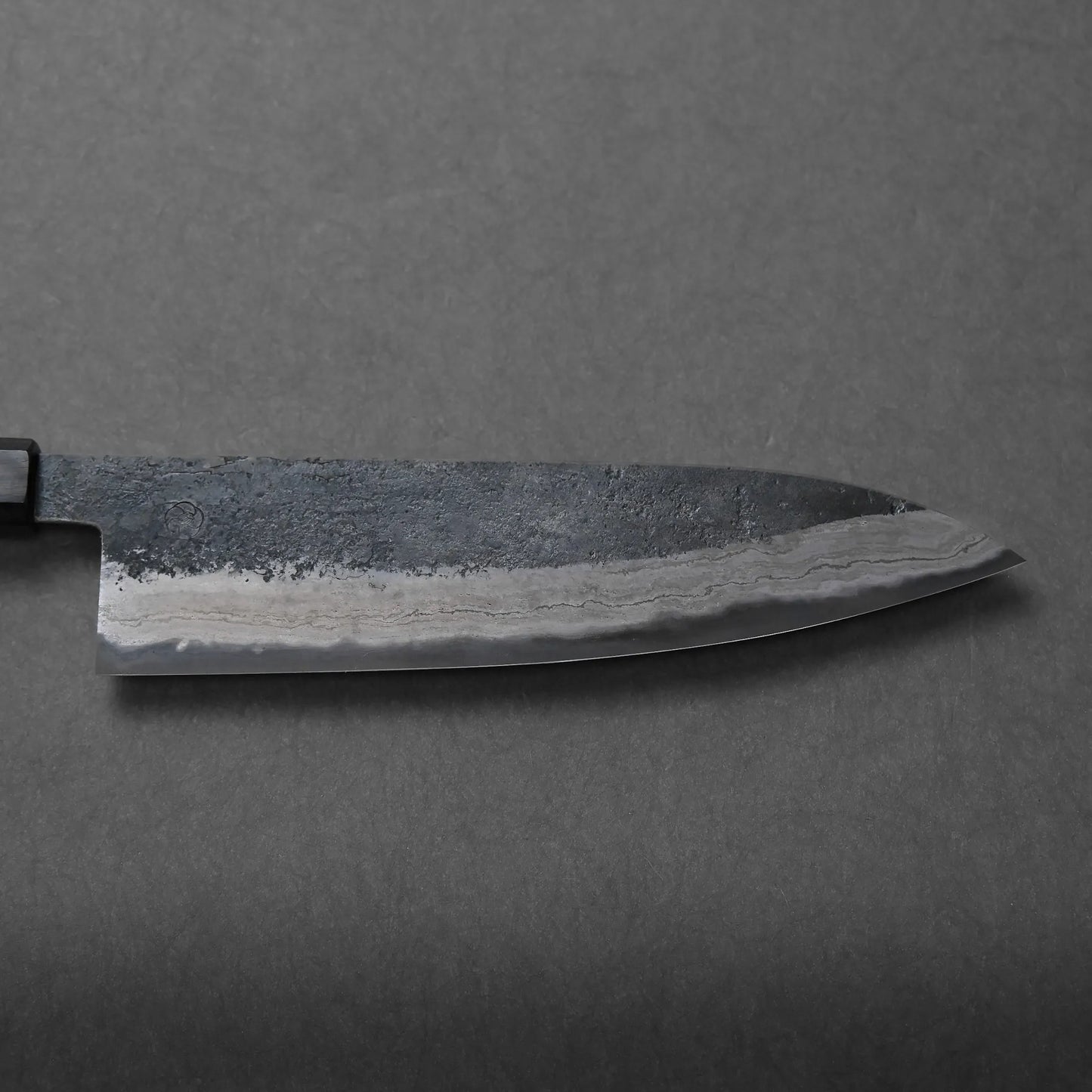 A close-up view of a chef's knife with a dark, textured blade featuring a distinct Damascus wavy pattern along the edge, resting on a plain gray surface.