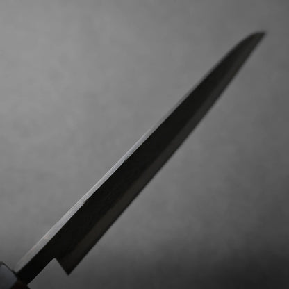 A close-up of a knife blade photographed against a plain, blurred gray background, with the blade's edge in focus and the handle partially visible at the lower left corner.