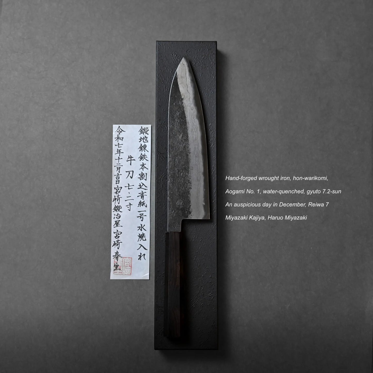 A hand-forged Japanese gyuto knife with a dark wooden handle rests on a black rectangular stand. To the left is a paper with Japanese calligraphy and a small red stamp, showcasing traditional craftsmanship. Text appears in the lower right corner.