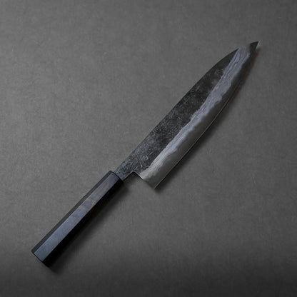 A Miyazaki kurouchi rentetsu aogami#1 gyuto 250mm with a dark, rectangular handle and a polished, patterned blade rests on a gray surface, showcasing elegant craftsmanship.