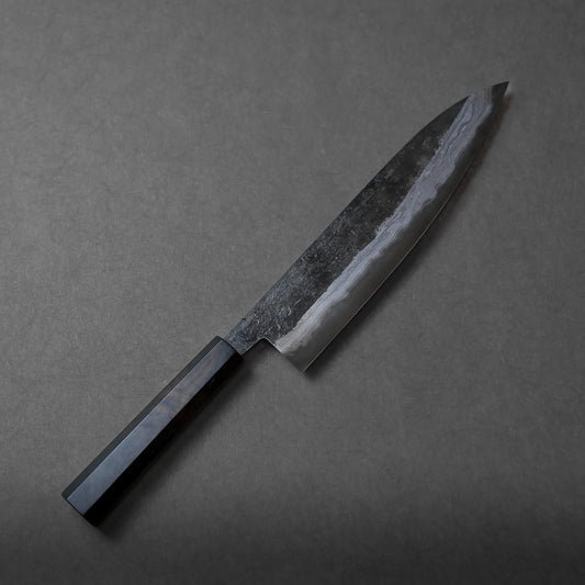A Miyazaki kurouchi rentetsu aogami#1 gyuto 250mm with a dark, rectangular handle and a polished, patterned blade rests on a gray surface, showcasing elegant craftsmanship.