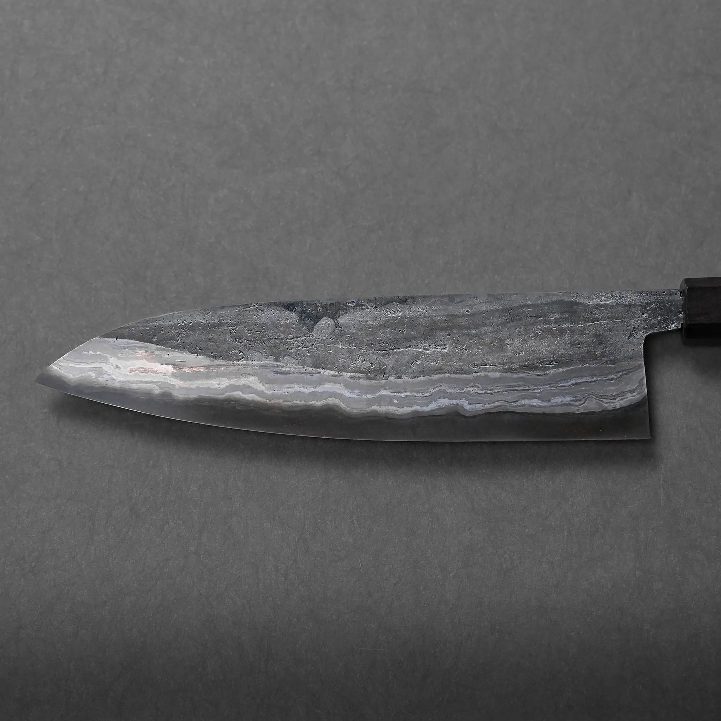 A close-up of a Japanese kitchen knife featuring a patterned, wavy Damascus steel texture on the blade, photographed against a plain, dark gray background.