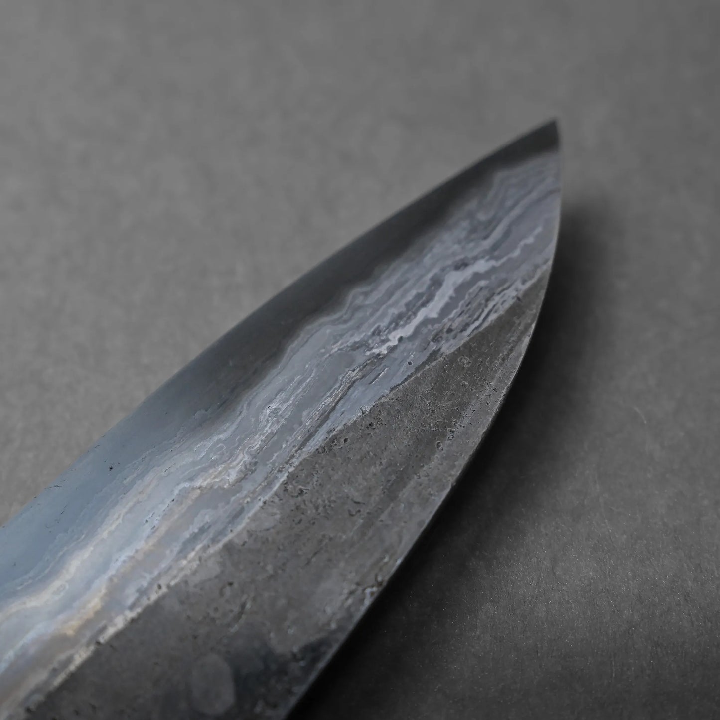 Close-up of the tip of a knife blade with visible layered patterns in the metal, resting on a plain gray surface. The striking Damascus design highlights the craftsmanship.