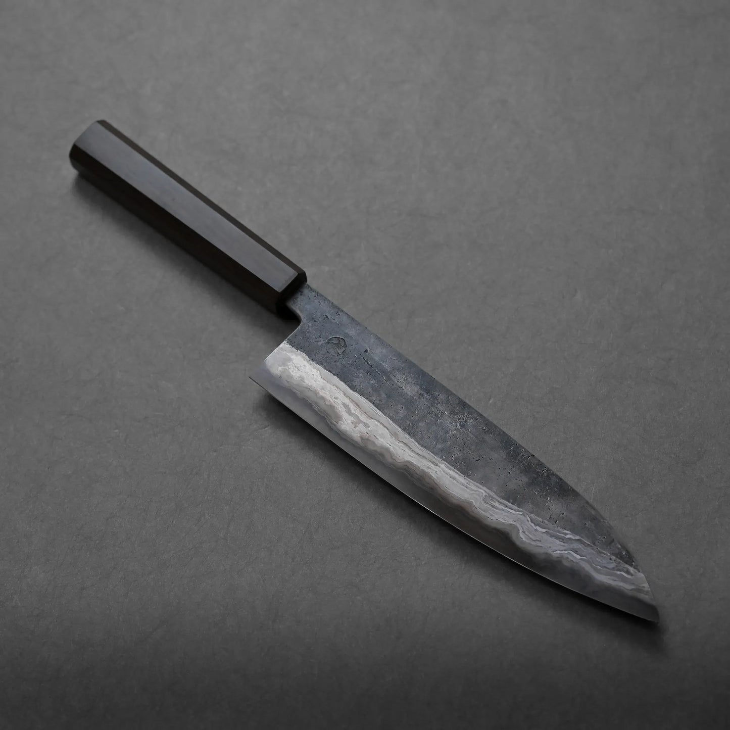 A chef’s knife with a dark wooden handle and a patterned blade rests on a gray surface, showcasing its elegant craftsmanship.