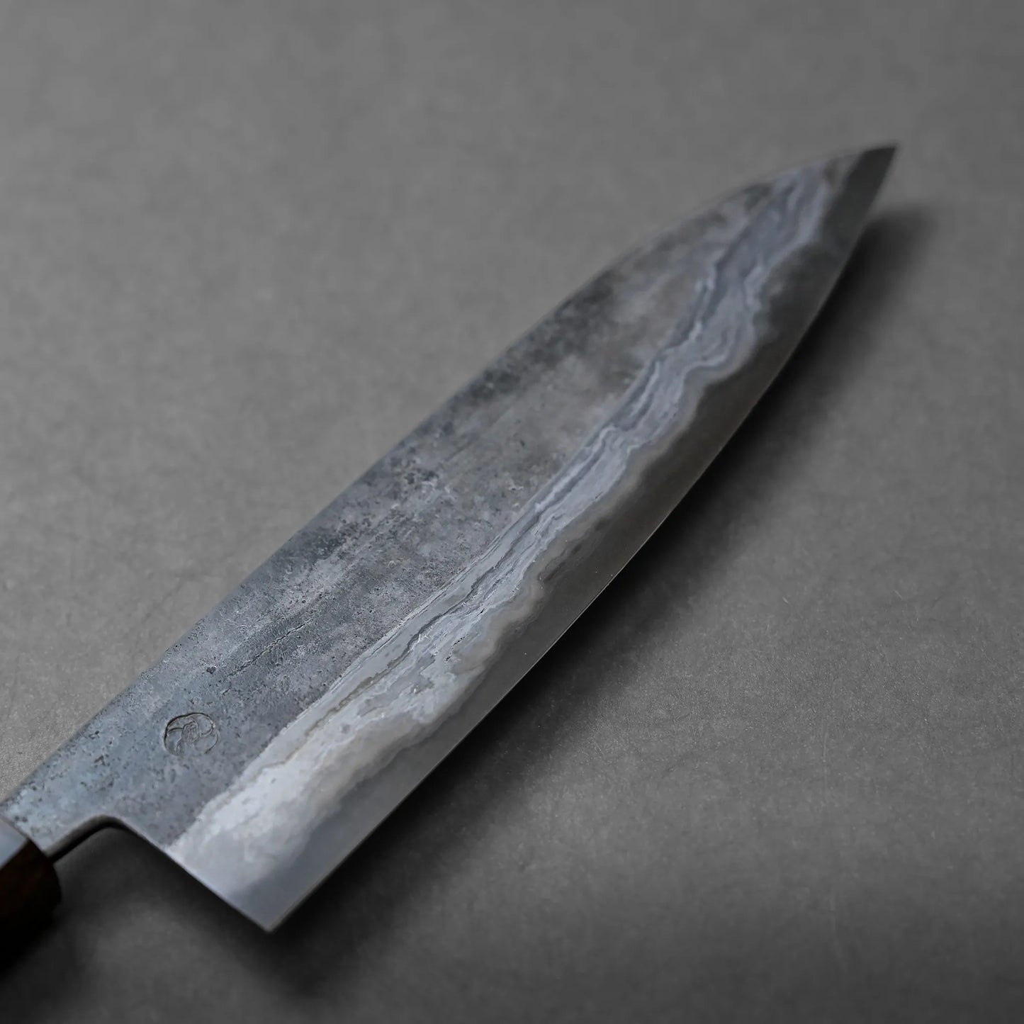 A close-up of a Japanese chef’s knife featuring a striking wavy pattern on its blade, resting on a plain gray surface.