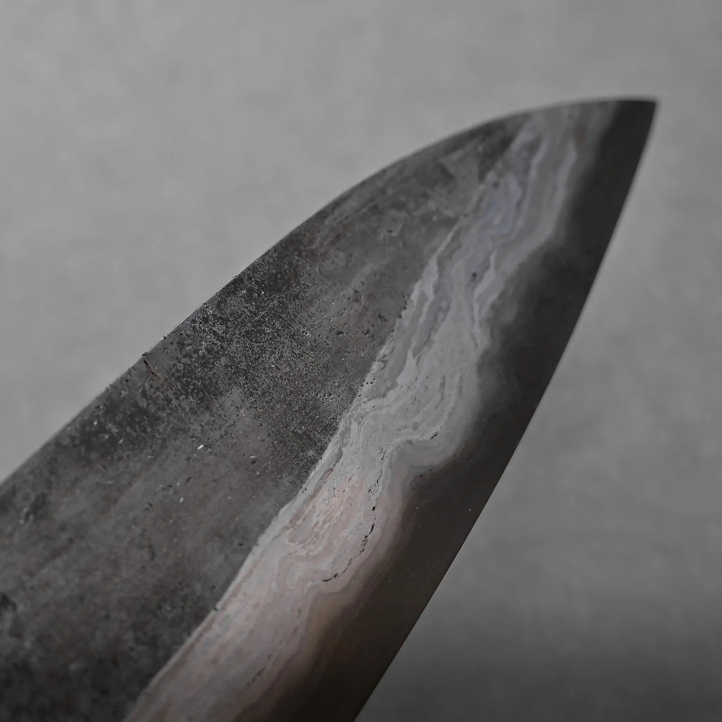 Close-up of the tip of a Damascus steel knife blade, featuring distinctive layered patterns and a matte, slightly textured surface against a plain gray background, highlighting its unique craftsmanship.