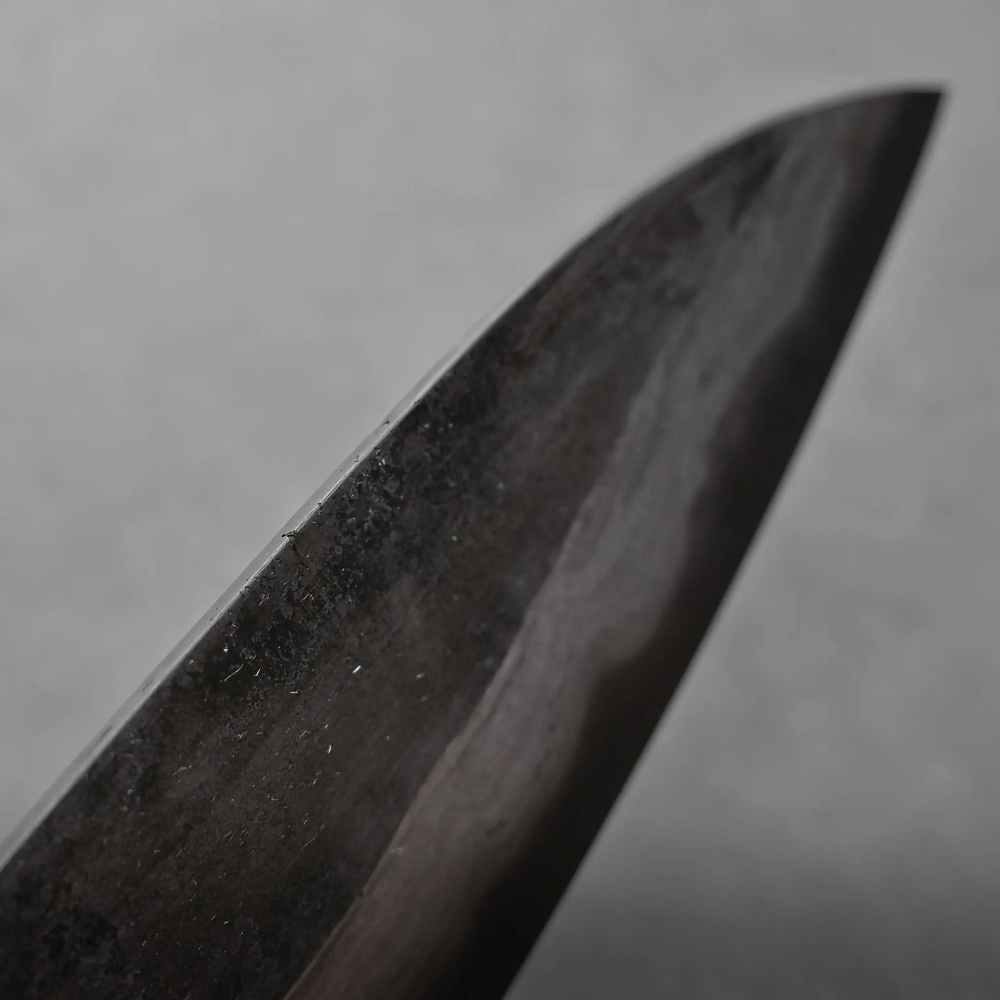 Close-up view of the dark, honed edge of a knife blade against a soft, gray background. The surface of the blade displays subtle texture and reflections.