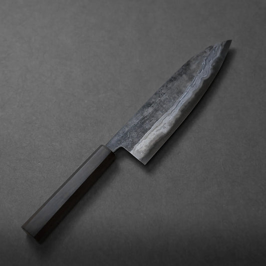 A Miyazaki kurouchi rentetsu aogami#2 gyuto 215mm with a dark wooden handle and a patterned steel blade, featuring subtle craftsmanship, rests on a dark gray surface.