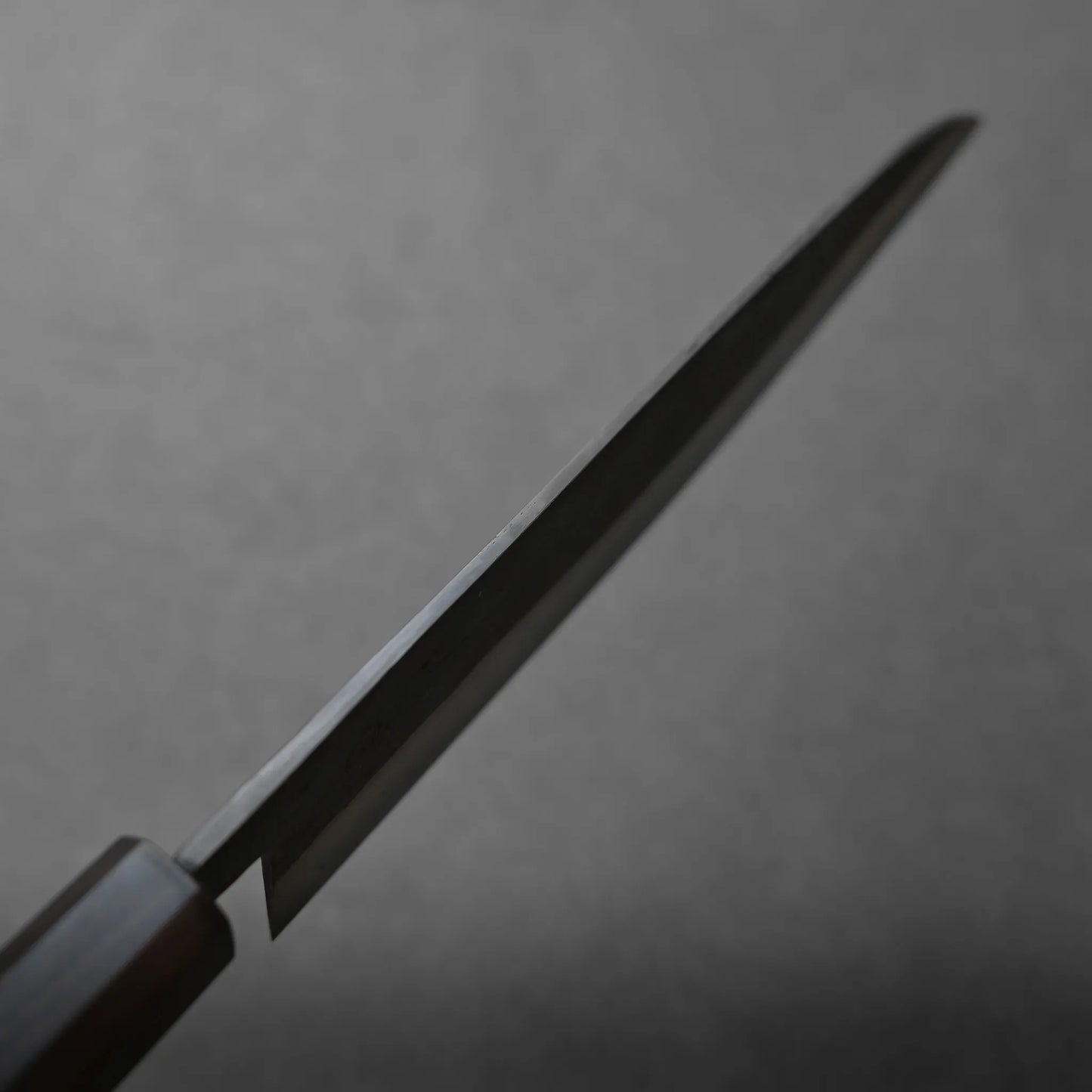 A close-up photo of a kitchen knife blade angled upward against a plain, blurred gray background. The handle appears partially visible on the left side of the image.