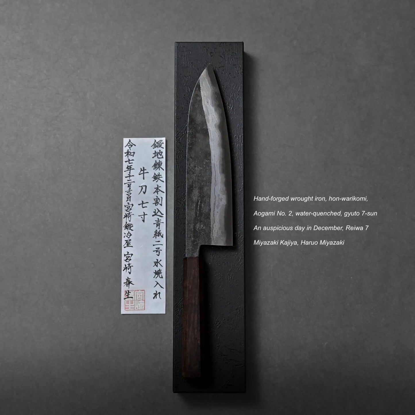 A hand-forged kitchen knife with a dark wooden handle is displayed on a black rectangular base, accompanied by Japanese calligraphy and an English description highlighting the craftsmanship behind the knife.