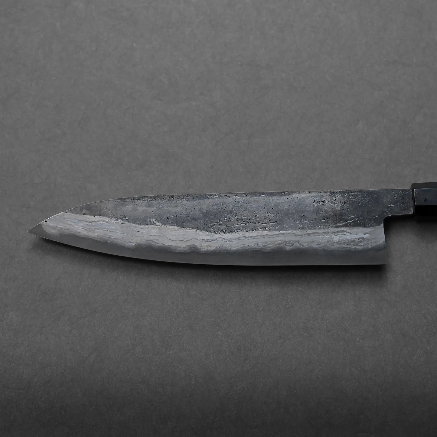 A close-up view of a Japanese-style chef’s knife with a dark blade and visible wavy pattern, resting on a gray textured surface, highlights its traditional craftsmanship.