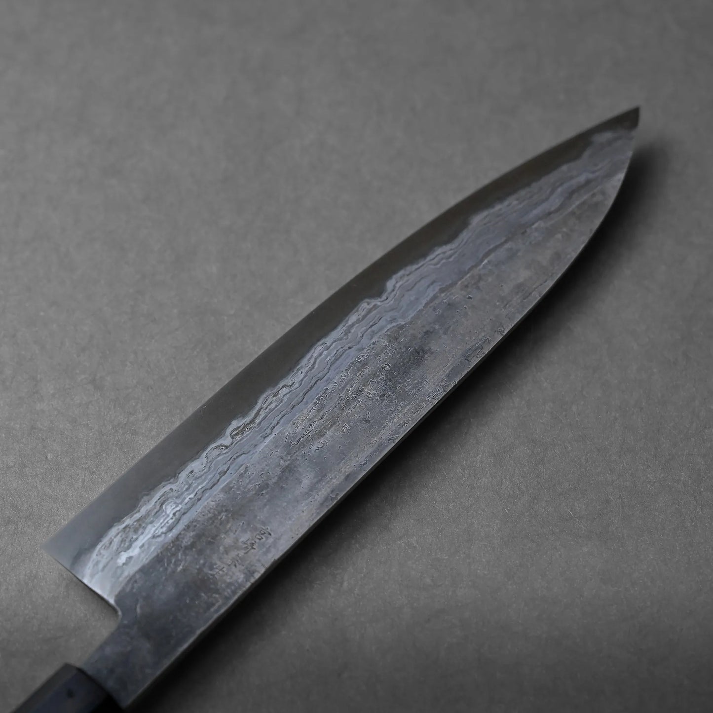 A close-up of a chef’s knife with a dark, patterned blade and a pointed tip, positioned on a plain grey surface. The blade displays wavy lines and striking texture characteristic of Damascus steel craftsmanship.