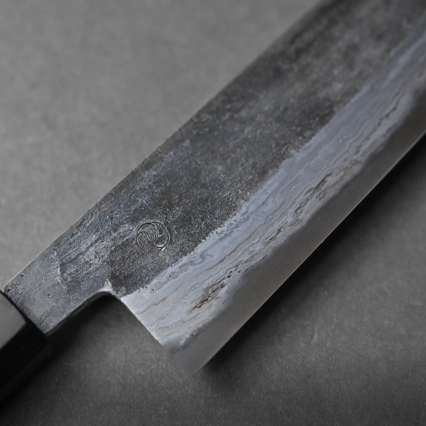 Close-up of a Japanese kitchen knife with a dark, textured blade and visible Damascus patterns near the edge, resting on a gray surface.