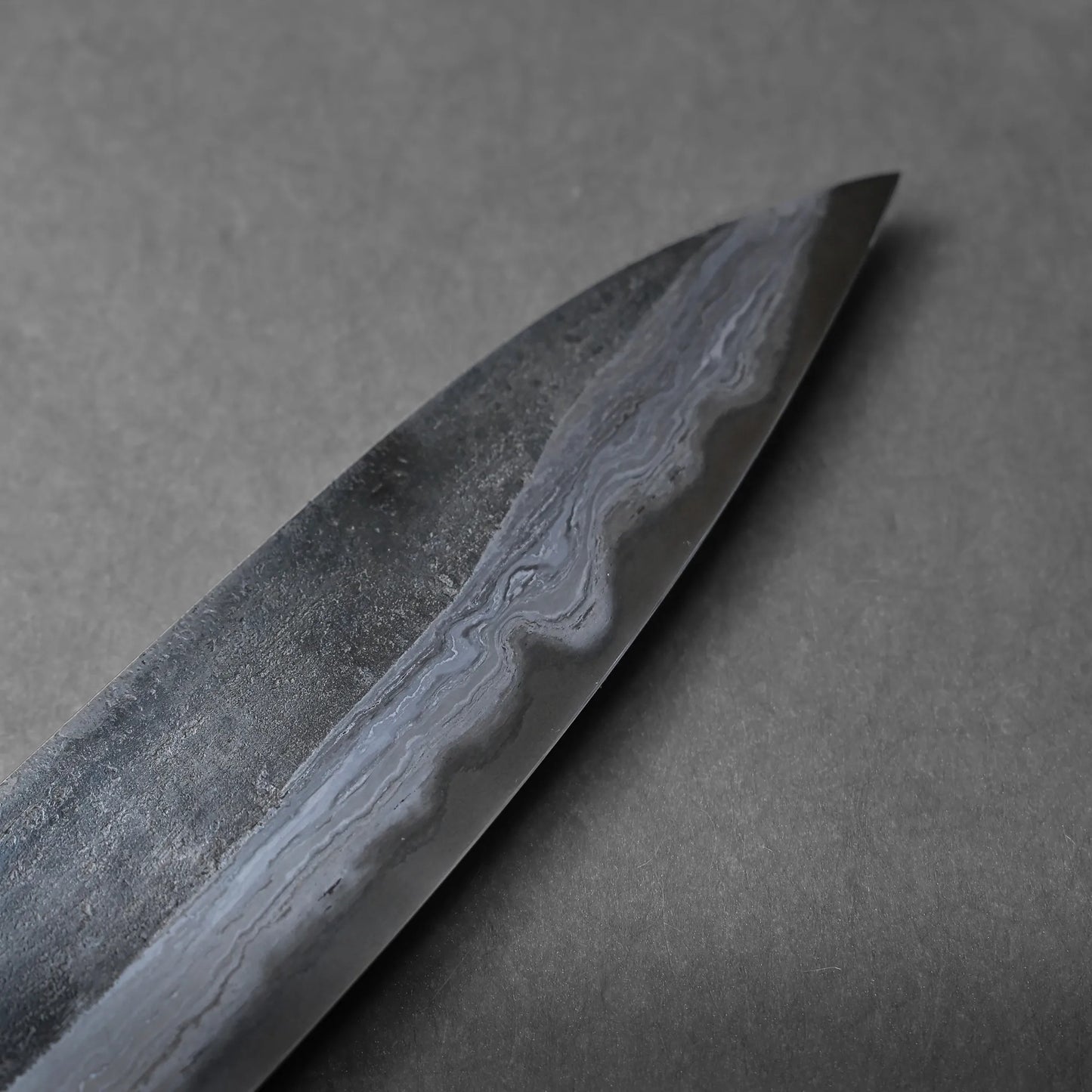 Close-up of a Damascus steel knife blade highlighting its wavy, layered pattern on the metal surface, photographed against a plain gray background.