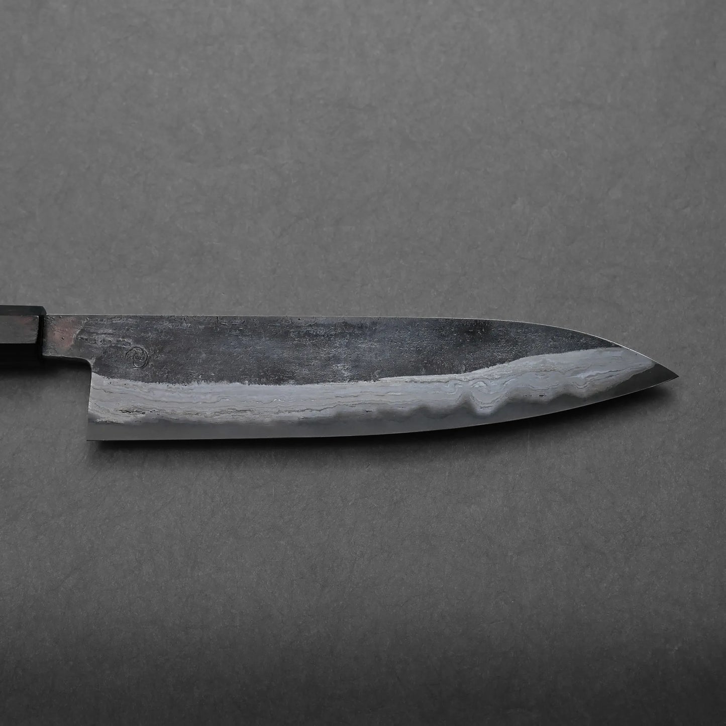 A close-up of a Japanese-style kitchen knife with a dark blade and visible layered patterns rests on a gray textured surface, highlighting its traditional craftsmanship. The knife is shown horizontally with the handle partially visible.