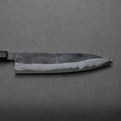 A close-up of a Japanese-style kitchen knife with a dark blade and visible layered patterns rests on a gray textured surface, highlighting its traditional craftsmanship. The knife is shown horizontally with the handle partially visible.