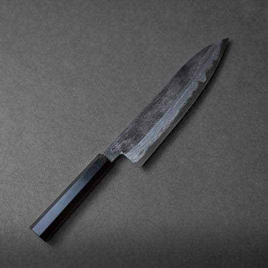 A Miyazaki kurouchi rentetsu aogami#2 gyuto 240mm with a dark, rectangular handle and a slightly curved, patterned blade rests on a dark grey surface, showcasing traditional craftsmanship.