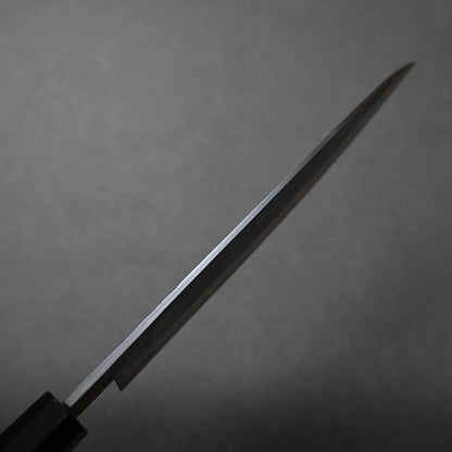 A close-up view of the blade of a stainless steel knife, positioned at an angle against a plain grey background. The blade is thin and reflective, with the handle partially visible in the lower left corner.
