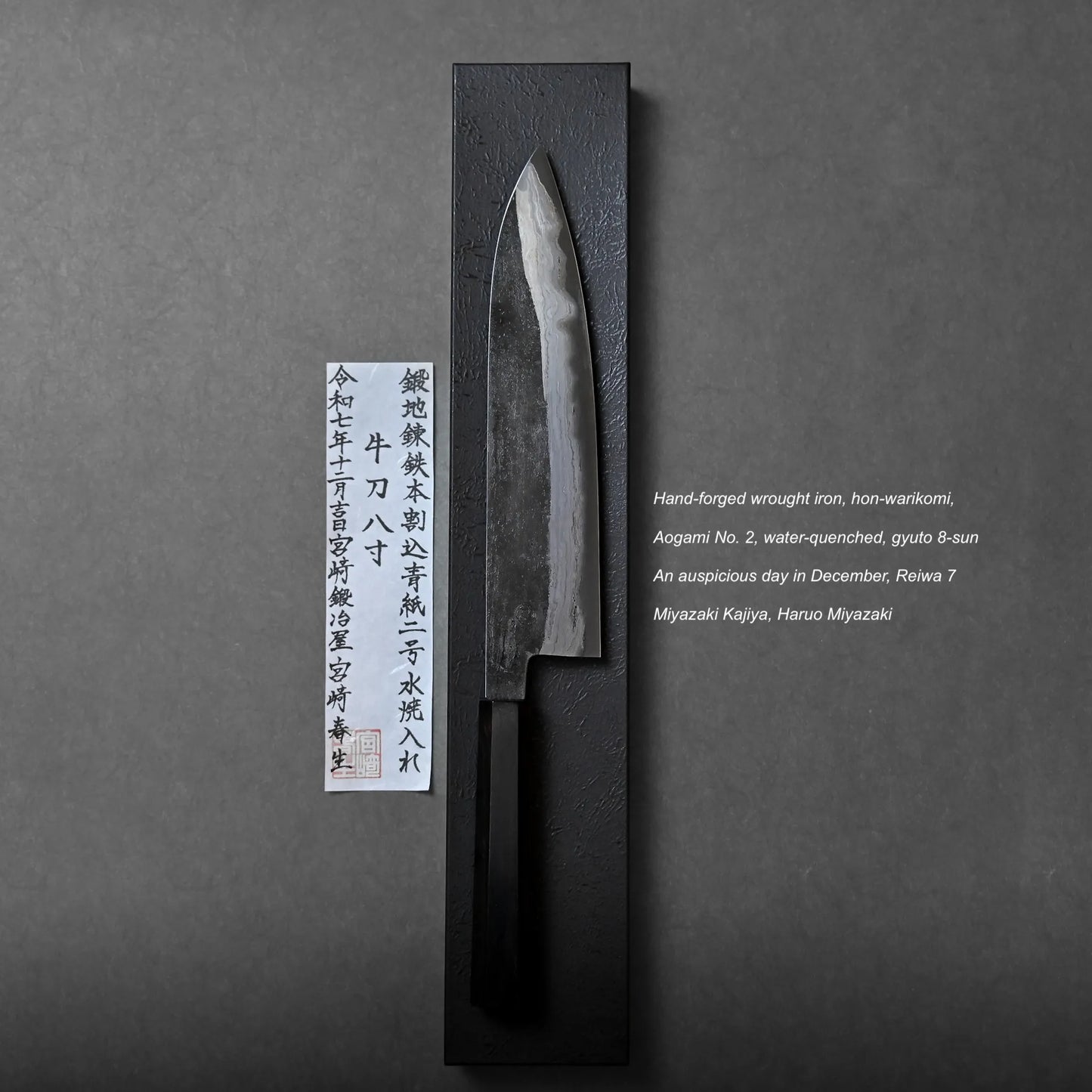 A hand-forged Japanese knife with a dark handle rests on a black rectangular stand beside a vertical paper label with Japanese text. English product details and makers’ names appear on the right.