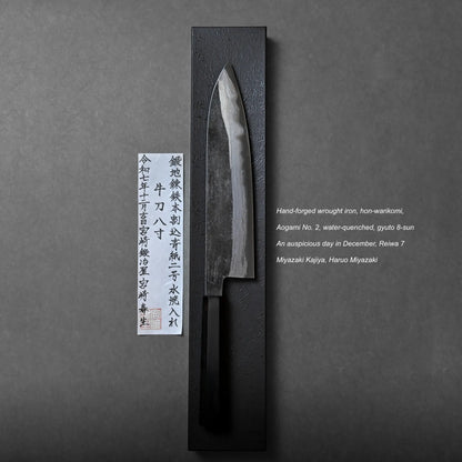 A hand-forged Japanese knife with a dark handle rests on a black rectangular stand beside a vertical paper label with Japanese text. English product details and makers’ names appear on the right.