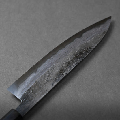 A close-up of a Japanese kitchen knife blade with a wavy Damascus pattern and textured surface, placed on a plain grey background.