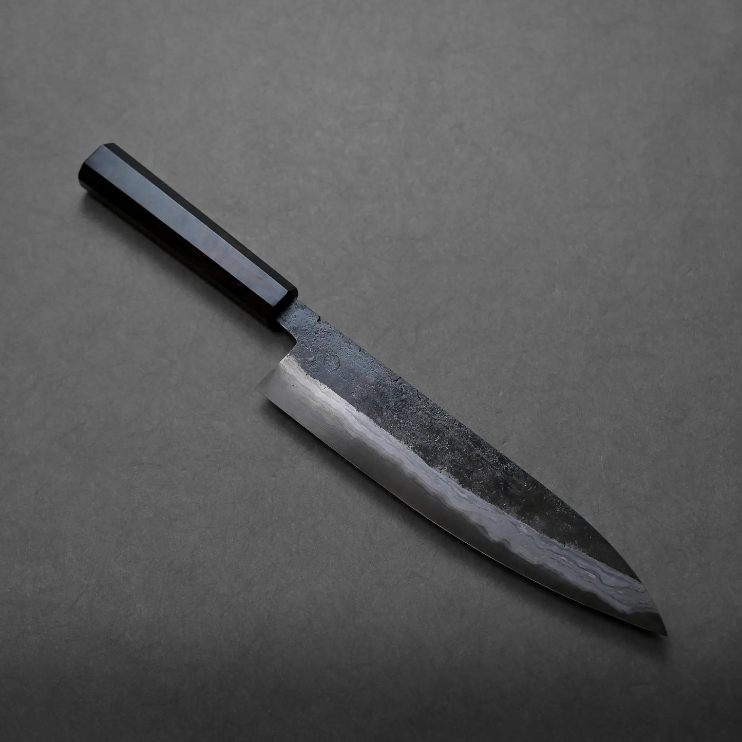 A chef’s knife featuring a dark wooden handle and a polished, slightly reflective blade rests on a plain dark grey surface. The elegant finish complements the craftsmanship.