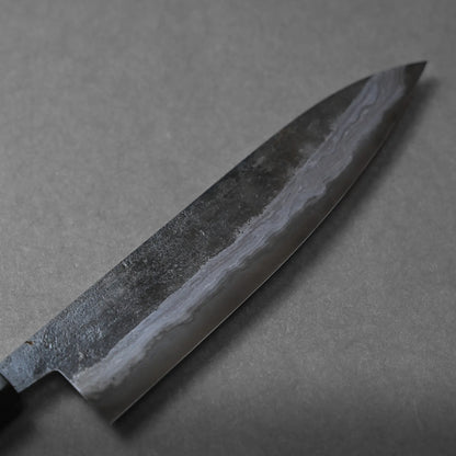 Close-up of a Japanese chef’s knife with a dark, patterned blade and a slight sheen, resting on a textured gray surface. The blade reveals distinct layers, characteristic of Damascus steel craftsmanship.