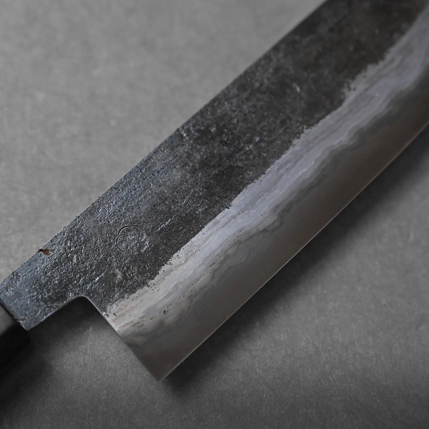 A close-up view of a Japanese kitchen knife blade with a wavy hamon line and a dark, textured upper portion, crafted using traditional methods, lying on a gray surface.