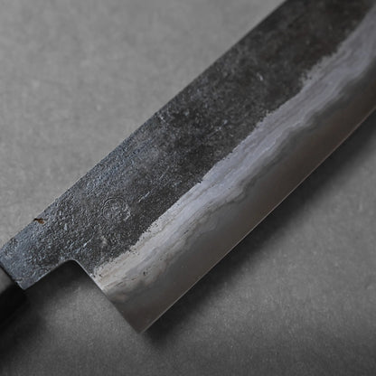 A close-up view of a Japanese kitchen knife blade with a wavy hamon line and a dark, textured upper portion, crafted using traditional methods, lying on a gray surface.